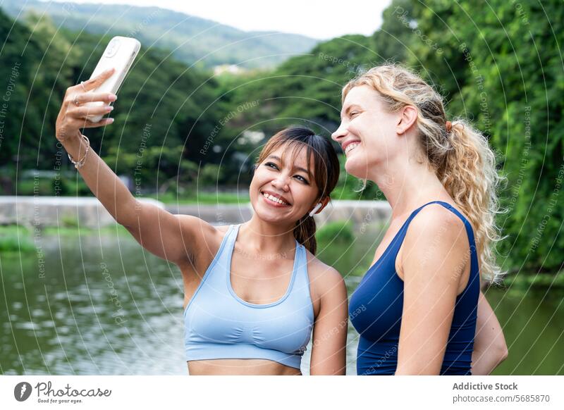 Glückliche Frauen nehmen Selfie in der Nähe von Teich heiter Sportlerinnen Smartphone Selbstportrait Pause Sportkleidung Berge u. Gebirge Training Freund