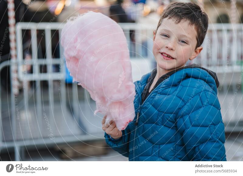 Fröhlicher Junge in einer blauen Jacke, der in die Kamera schaut, während er in einem Vergnügungspark riesige Zuckerwatte hält Vergnügen Park Leckerbissen süß