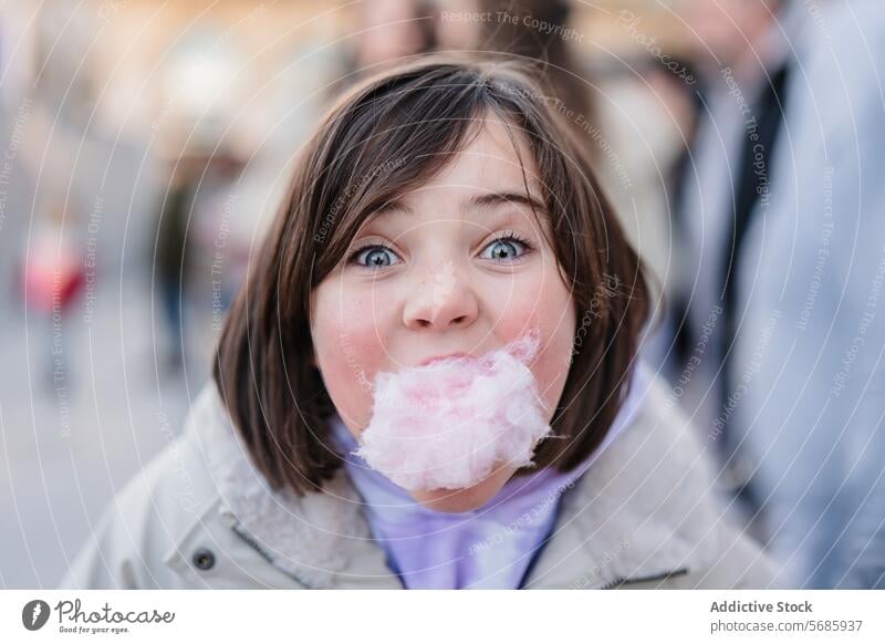 Nahaufnahme eines aufgeregten Mädchens mit großen Augen, das in die Kamera schaut, während es einen Bissen Zuckerwatte auf einem unscharfen Straßenhintergrund genießt