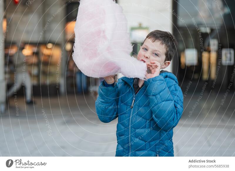 Ein Junge in einer blauen Jacke schaut in die Kamera und beißt in eine flauschige rosa Zuckerwatte beißend fluffig Geschmack Snack Konfekt süß zuckerhaltig