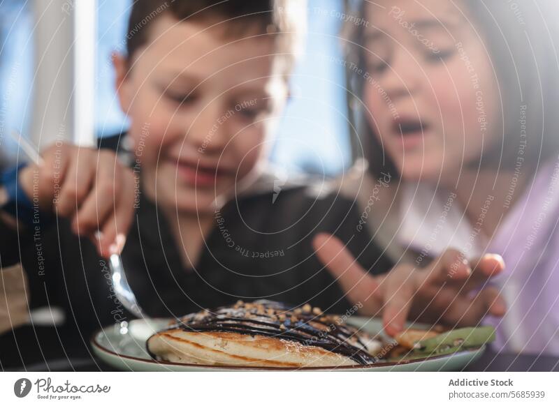 Zwei Kinder genießen eine gemeinsame Mahlzeit, ein Junge hält eine Gabel über einen Pfannkuchen mit Sirup, ein Mädchen zeigt auf ihn. Belag Zeigen genießend