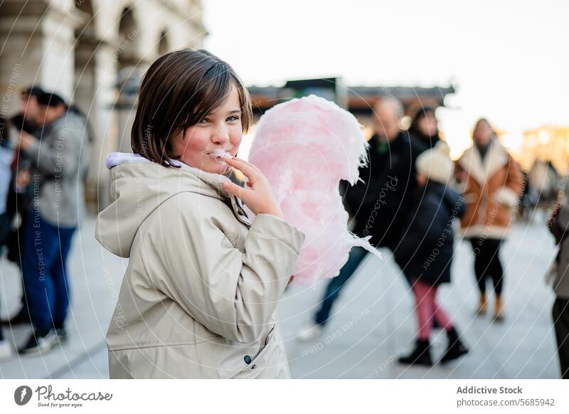 Seitenansicht eines lächelnden Mädchens in einem beigen Mantel, das spielerisch Zuckerwatte auf einer belebten Straße isst genießend Essen Lächeln beschäftigt