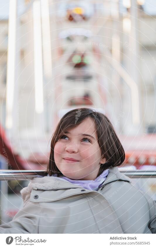 Ein Mädchen in einem beigen Mantel mit verträumtem Gesichtsausdruck schaut auf, während es vor einem Riesenrad sitzt Ausdruck Aussehen nach oben Vergnügen Park