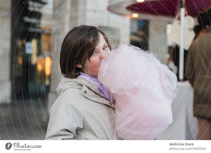 Ein Mädchen mit einer beigen Jacke schaut in die Kamera, während es auf einer Straße in der Stadt eine große rosa Zuckerwatte isst Essen Großstadt im Freien süß