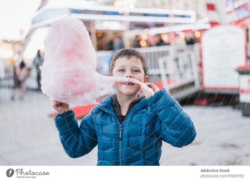 Glücklicher Junge in einem blauen Mantel, der in die Kamera schaut, während er mit einem großen Stück Zuckerwatte auf einem Rummelplatz posiert posierend