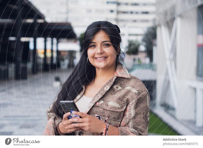 Glückliche Frau mit Smartphone in der Nähe einer Glaswand in der Stadt selbstbewusst Jeanshose Turnschuh Lächeln Großstadt Straße modern Stil urban benutzend