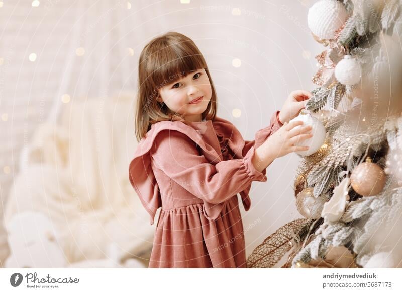 Kleine adorable Mädchen in weichen rosa Kleid hängt weiß glänzende Kugel auf Weihnachtsbaum mit Licht. Kind schmücken Xmas Baum in schönen Wohnzimmer. Glückliche Familie Urlaub Neujahr Konzept.
