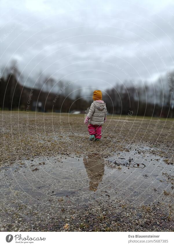 Warm angezogenes Kleinkind geht von einer Pfütze weg Winter Pfützenspiegelung Kind Regen Wetter nass trist
