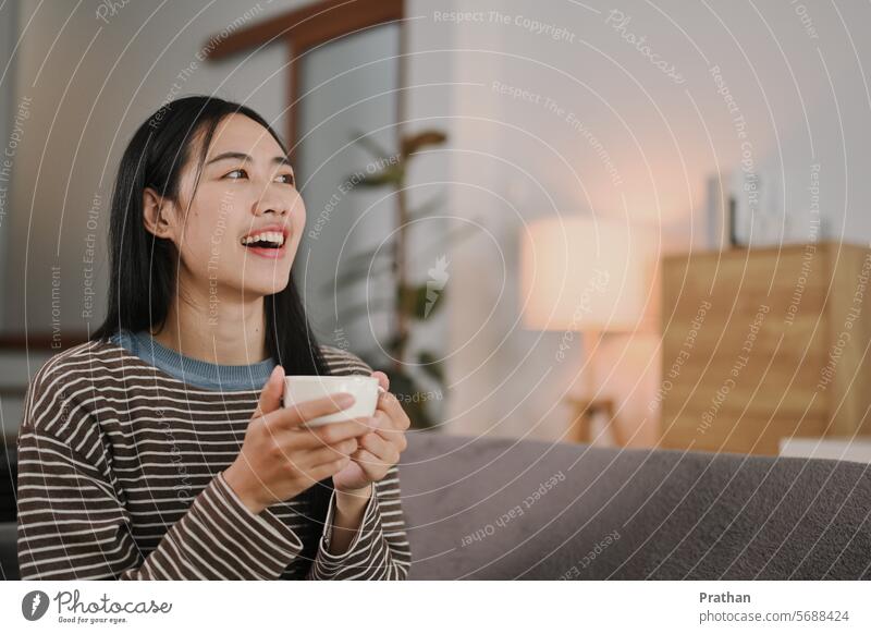 Lächelnde junge Frau, die sich mit einem Kaffee auf dem Sofa entspannt, wegschaut und über etwas nachdenkt Kaffeetasse Getränk Morgen entspannend Wohnzimmer