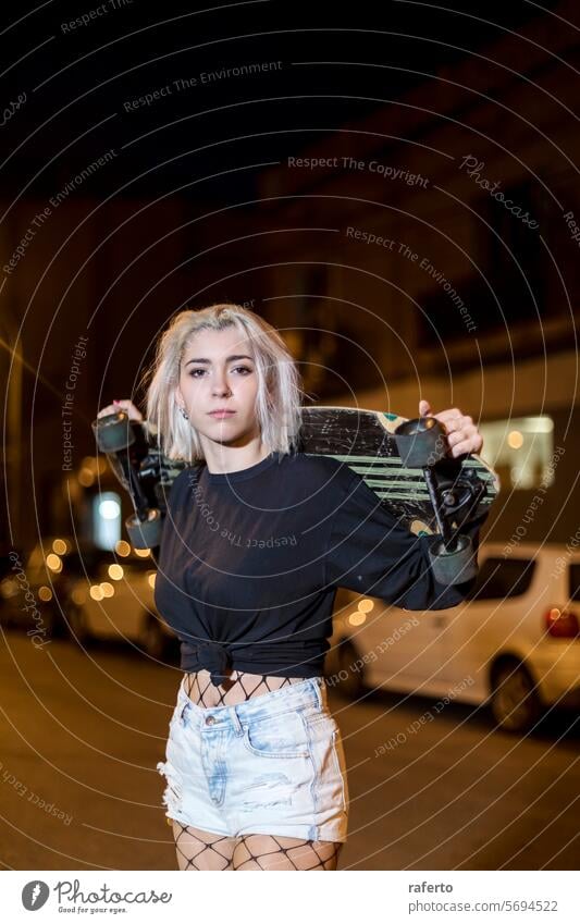 Frau mit Kamera in Shorts, die nachts auf der Straße steht und ein Skateboard hält brünett Selbstvertrauen cool modisch Hipster Erholung Haut Jugend blond Nacht