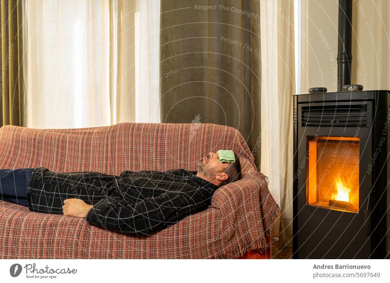 Ein kranker spanischer Mann liegt mit einem Bademantel und einem Tuch auf der Stirn in der Wärme eines Pelletofens auf der Couch. männlich Sofa Fieber