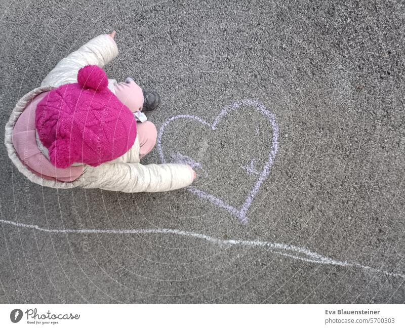 Kind mit pinker Mütze bemalt Herz auf Straße mit Straßenmalkreide Liebe malen Straßenkreide Strassenmalerei Kreide Kinderzeichnung Asphalt