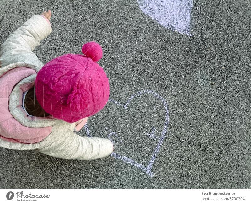 Kind mit pinker Mütze bemalt Herz auf Straße mit Straßenmalkreide Liebe malen Straßenkreide Strassenmalerei Kreide Kinderzeichnung Asphalt