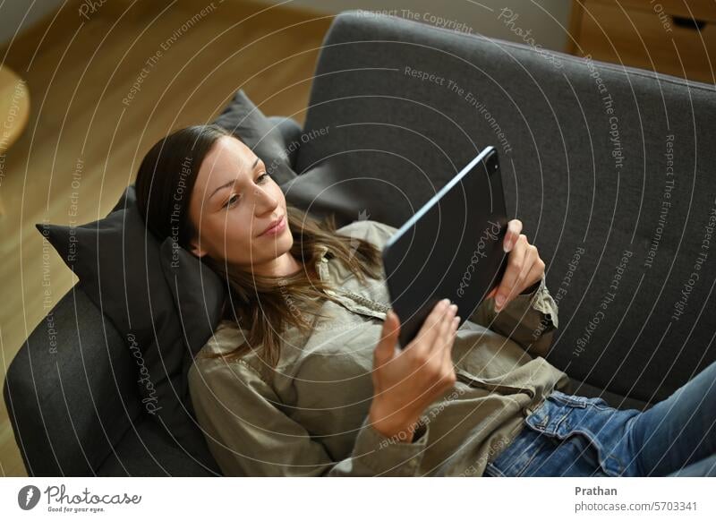 Entspannte junge Frau, die auf einer Couch im Wohnzimmer liegt und ein Video auf einem digitalen Tablet ansieht digitales Tablett Technik & Technologie Winter