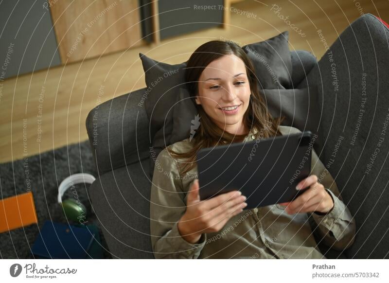 Lächelnde junge Frau, die auf der Couch liegt und im Internet surft oder ein Video auf einem digitalen Tablet ansieht digitales Tablett Technik & Technologie