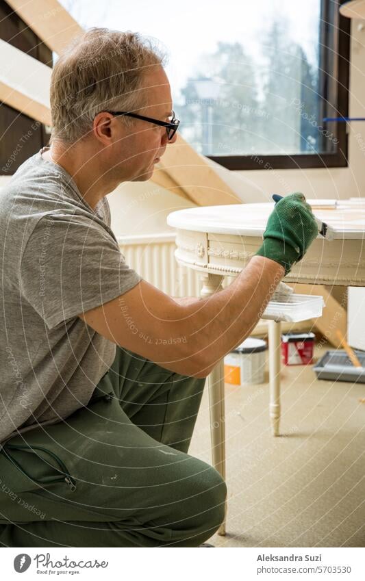 Middle-age Mann Malerei alten Holztisch mit Pinsel in weißer Farbe. Möbelrestaurierung, DIY-Reparatur, Selbstreparatur und Heimwerken. Recycling und nachhaltiger Lebensstil
