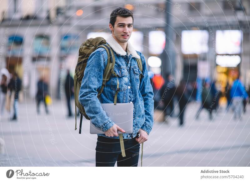 Ein selbstbewusster junger Mann mit einem Laptop steht auf einem belebten Platz in Madrid Quadrat professionell Rucksack Jeansstoff Jacke urban Stadtbild