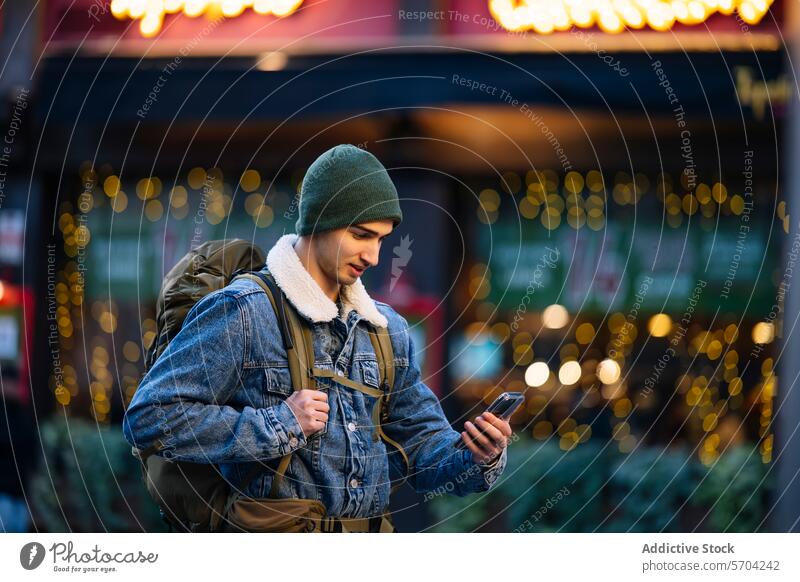 Junger Mann mit Mütze und Jeansjacke, der sein Telefon benutzt, mit funkelnden Stadtlichtern im Hintergrund jung Beanie Jeansstoff Jacke Großstadt Lichter Bokeh