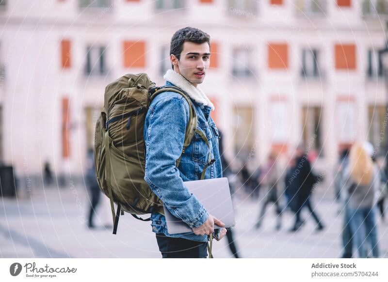 Seitenansicht eines jungen Mannes mit Rucksack und Laptop, der auf einer belebten Straße in Madrid zurückblickt technikbegeistert Reisender Jeansstoff Jacke
