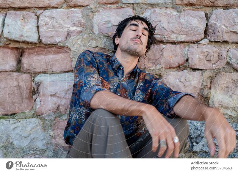 Nachdenklicher Mann sitzt an einer rustikalen Steinmauer beschaulich Sitzen Steinwand Gedanke jung Ausdruck besinnlich gealtert in Gedanken versunken lässig
