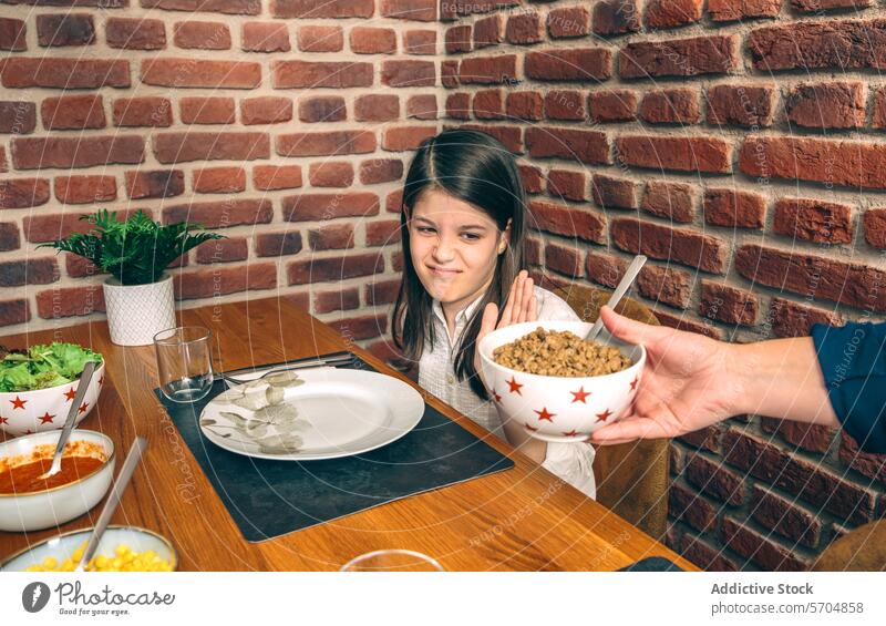 Ein junges Mädchen schaut skeptisch auf eine Schüssel mit Essen, die ihr am Esstisch der Familie vor einem Backsteinmauer-Hintergrund angeboten wird