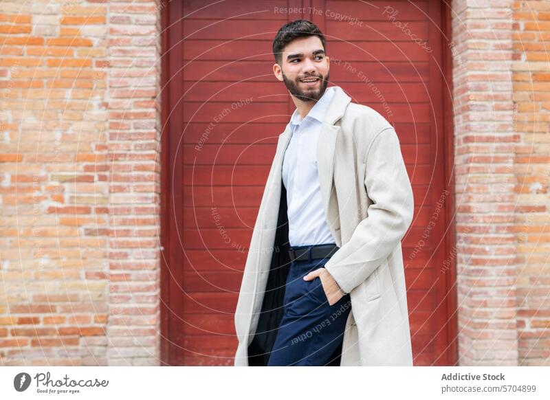 Ein stilvoller junger Mann in Geschäftskleidung lächelt selbstbewusst, während er vor einem roten Türhintergrund steht und in seinem beigen Mantel und der marineblauen Hose ein Gefühl von lässiger Professionalität ausstrahlt.