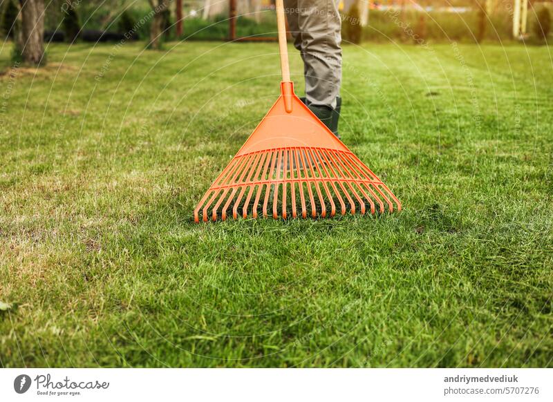 Männlicher Gärtner sammelt geschnittenes Gras mit orangefarbener Plastikharke nach einem Rasenmäher, arbeitet im Hinterhof des Hauses. Mann kümmert sich um den Rasen. Konzept der Hausarbeit, Gartenarbeit und Landleben, Gartengeräte.