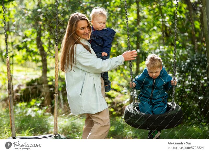 Die Mutter mit zwei Kindern vergnügt sich auf dem Spielplatz. Die Mutter schaukelt ihre Kinder auf der Schaukel. aktiv Aktivität Baby Junge Pflege Kaukasier