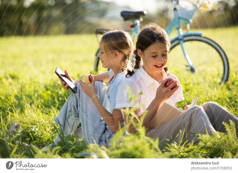 Zwei Mädchen verbringen gemeinsam Zeit auf dem grünen Rasen im Park, scrollen durch ihre Smartphones, lesen Bücher, essen Äpfel und genießen den Sommer und die Ferien