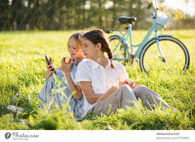 Zwei Mädchen verbringen gemeinsam Zeit auf dem grünen Rasen im Park, scrollen durch ihre Smartphones, lesen Bücher, essen Äpfel und genießen den Sommer und die Ferien
