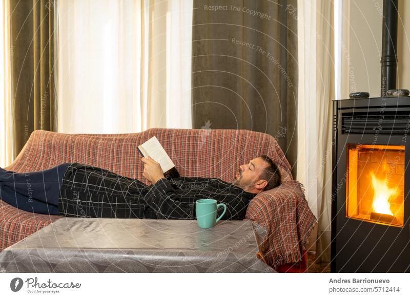 Hispanischer Mann mit Bart in den 40ern liegt auf der Couch und liest ein Buch, er hat es sich mit der Wärme eines Pelletofens sehr bequem gemacht Herd ifestyle
