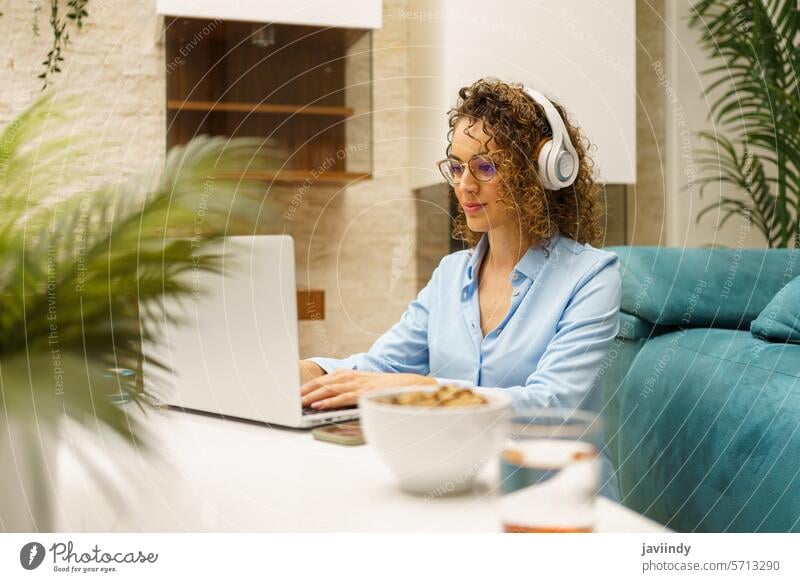 Positive Frau mit Kopfhörern, die im Wohnzimmer auf einem Laptop tippt freiberuflich Tippen abgelegen Arbeit heimwärts positiv Projekt online Job Internet
