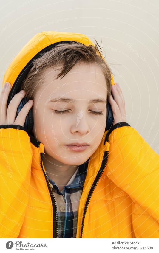 Ein nachdenklicher Junge in einer auffälligen gelben Jacke genießt Musik über seine Kopfhörer vor einem weichen, neutralen Hintergrund hören beschaulich