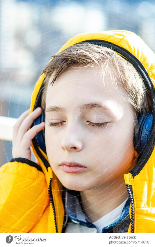 Ein gelassener Junge mit geschlossenen Augen genießt Musik über schwarze Kopfhörer und trägt einen leuchtend gelben Kapuzenmantel Mantel hören Gelassenheit