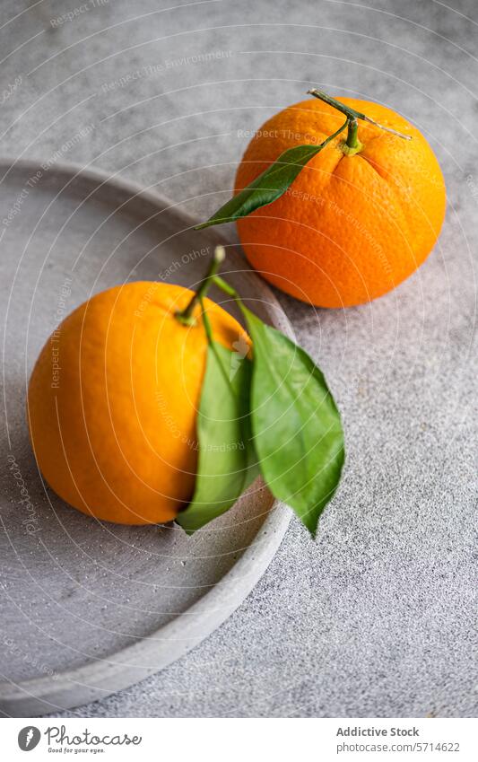 Nahaufnahme von zwei reifen Orangen mit Stielen und einem grünen Blatt auf einem strukturierten grauen Teller, mit einem Weichzeichner-Hintergrund orange Frucht