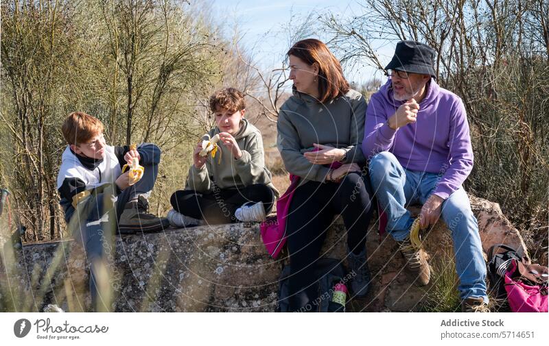 Eine Familie macht während einer Wanderung auf einer rustikalen Steinbank eine Snackpause, während zwei Kinder Bananen schälen und die Erwachsenen zusehen Pause