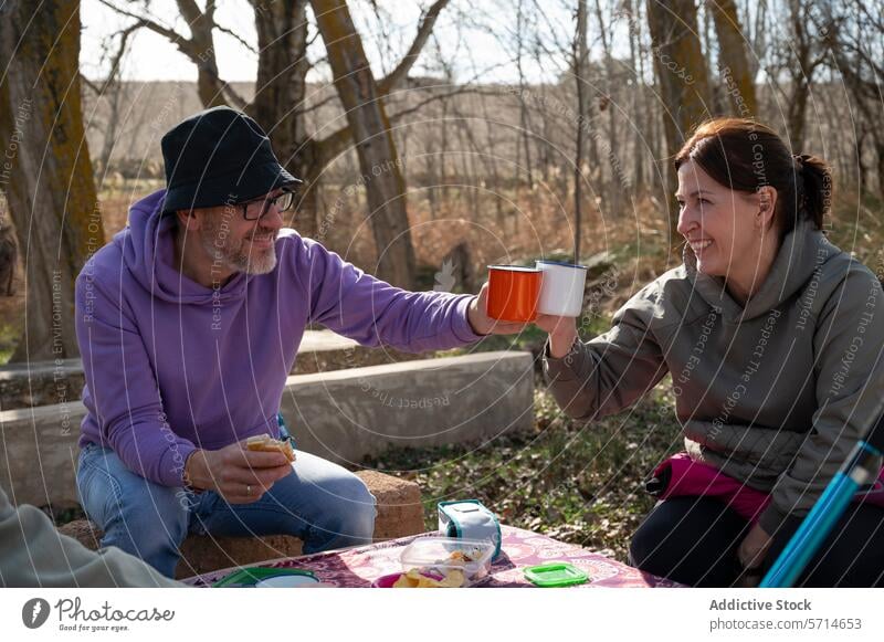Zwei Erwachsene genießen ein Picknick mit einem Toast und lächeln sich an, während sie an einem Tisch mit Snacks sitzen, umgeben von Bäumen Erwachsener