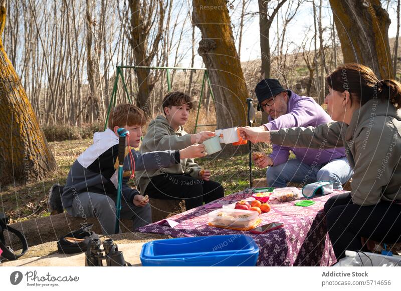 Eine Familie genießt ein Picknick im Wald und stößt mit Tassen an, während zwei Kinder und zwei Erwachsene um einen mit Tischtüchern gedeckten Tisch mit Essen sitzen