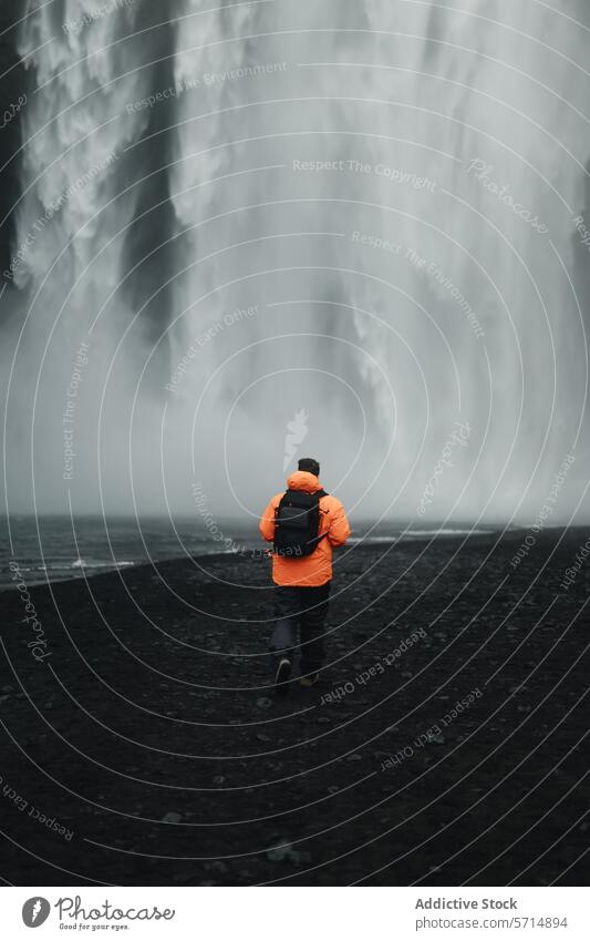 Ein Mann erkundet den majestätischen Wasserfall Skogafoss in Island männlich Abenteuer Entdecker reisen Natur vulkanisch Strand Wunder Rucksack orange Jacke