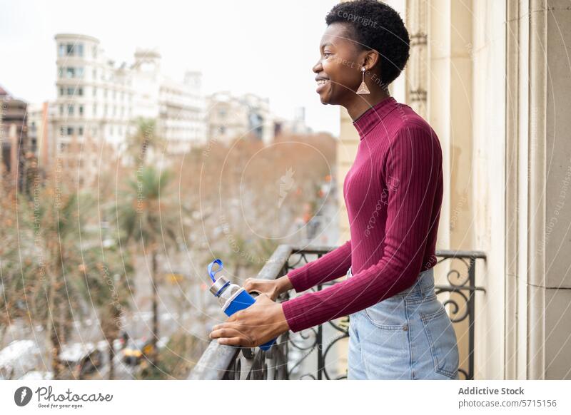 Lächelnde schwarze Frau mit Wasserflasche auf städtischem Balkon Stadtbild wiederverwendbar umweltfreundlich urban Glück Hydratation umgebungsbedingt im Freien