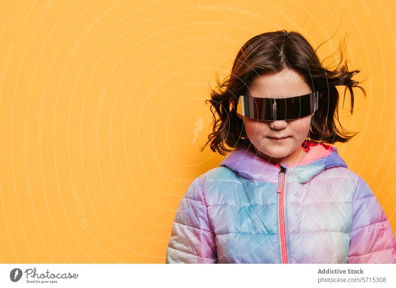 Selbstbewusstes junges Mädchen mit im Wind wehenden Haaren, einer übergroßen futuristischen Brille und einer bunten Jacke vor einem gelben Hintergrund