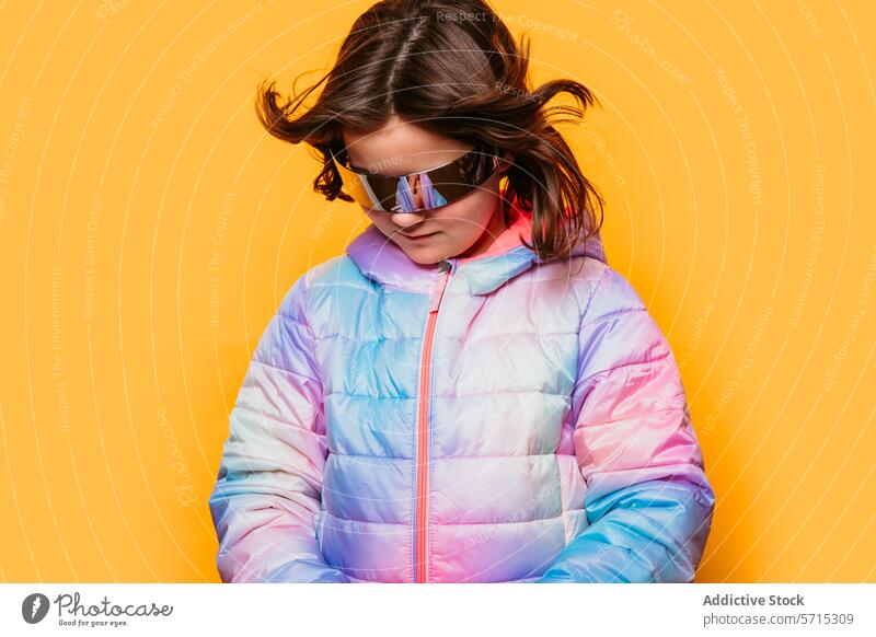 Junges Mädchen mit Wind in den Haaren, das eine futuristische Brille und eine pastellfarbene Pufferjacke auf einem orangefarbenen Hintergrund trägt herabsehend