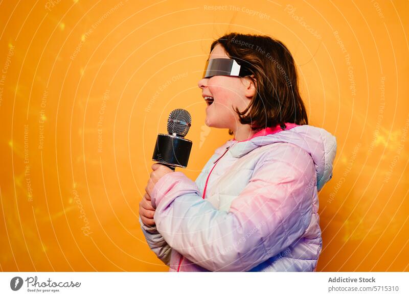Fröhliches Mädchen, das in ein Mikrofon singt, eine futuristische Brille und eine bunte Jacke trägt, auf gelbem Hintergrund Gesang gelber Hintergrund freudig
