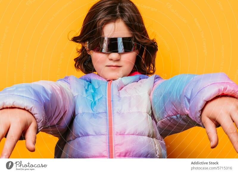 Selbstbewusstes Mädchen, das mit einer futuristischen Brille posiert und die Hände nach vorne streckt, in einer bunten pastellfarbenen Jacke vor einem orangefarbenen Hintergrund