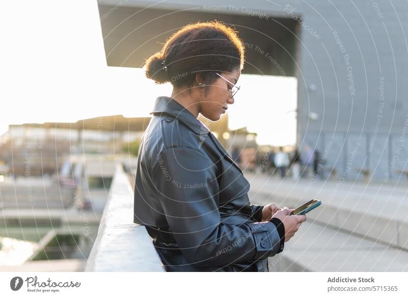 Eine Frau mit Wintermantel und Brille konzentriert sich auf ihr Smartphone, während sie bei Sonnenuntergang auf einer Stadtbrücke steht Brücke Großstadt Mantel