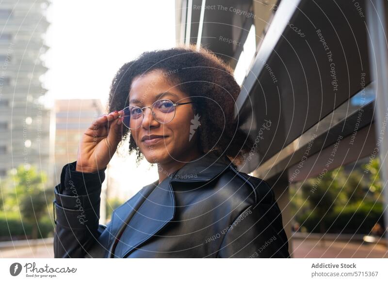 Selbstbewusste Frau in einem schicken schwarzen Mantel, die ihre Brille zurechtrückt, mit moderner Architektur im Hintergrund selbstbewusst Stil Mode urban