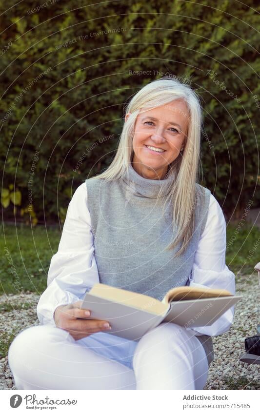 Lächelnde reife Frau, die im Freien sitzt und ein Buch liest, mit einer Kamera neben sich, die einen aktiven und engagierten Lebensstil zeigt lesen Lifestyle