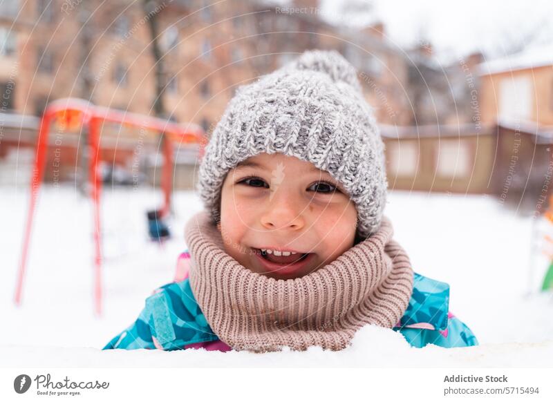 Lächelndes Kind mit Wollmütze und Schal, das einen verschneiten Tag auf dem Spielplatz genießt Schnee Winter Freude im Freien kalt heiter Spaß Saison