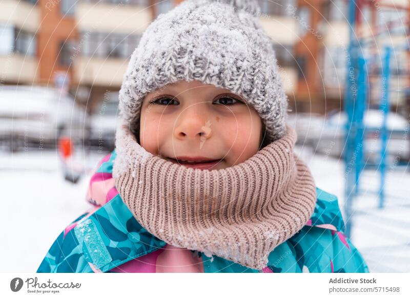 Lächelndes Kind mit gestrickter Mütze und Schal im Freien mit einem verschneiten Spielplatz im Hintergrund Strickmütze Winter heiter Fröhlichkeit kalt Wetter