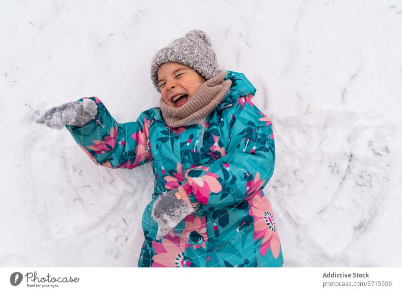 Freudig lachendes Kind, das einen Schneeengel macht, gekleidet in einen farbenfrohen Wintermantel und eine Mütze Lachen Hut Freude im Freien pulsierend kalt